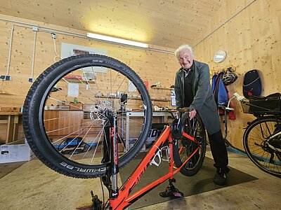 Ein älterer Mann steht in einer Werkstatt und repariert ein Fahrrad. Er hat graue Haare und trägt einen langen grünen Mantel. Das Fahrrad ist orange und hat ein großes Vorderrad. Um ihn herum ist eine Werkbank mit Werkzeugen und weiteren Fahrrädern.