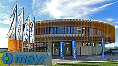Moderne Firmengebäude mit runder Architektur, umgeben von mehreren Fahnenmasten. Eine prominente Beschilderung mit dem Namen "mayr" ist im Vordergrund sichtbar. Der Himmel ist blau mit einigen Wolken. Die Umgebung ist grün und gepflegt.