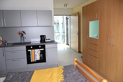 Ein modernes Zimmer mit einer Küchenzeile, die graue Schränke, einen Herd und einen Wasserkocher zeigt. Im Hintergrund ist eine Tür sichtbar, die zu einem Balkon führt. Links vom Bild liegt ein Bett mit einer gelben Decke.