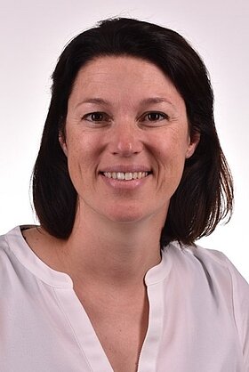 Porträt einer lächelnden Frau mit schulterlangen, dunklen Haaren, die ein weißes Oberteil trägt. Der Hintergrund ist hell und neutral, was den Fokus auf ihr freundliches Ausdruck lenkt.