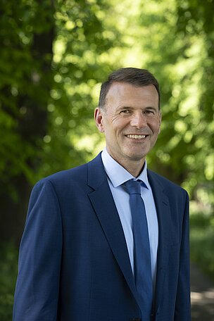 Ein Mann in einem blauen Anzug und Krawatte lächelt vor einem Hintergrund aus grünen Bäumen. Die Sonne scheint, und er steht in einer natürlichen Umgebung.