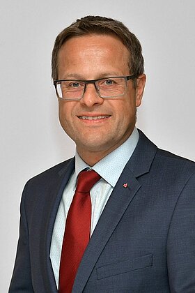Ein Mann in einem dunkelblauen Anzug mit roter Krawatte und einem hellblauen Hemd lächelt in die Kamera. Er trägt eine modische Brille und hat kurze, leicht gewellte Haare. Der Hintergrund ist einfarbig und neutral.