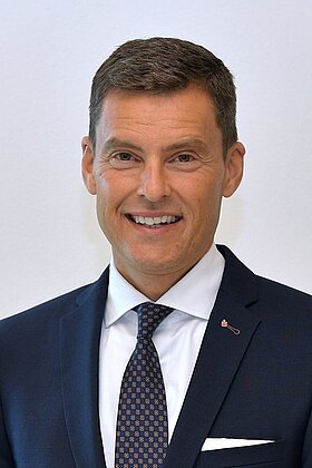 Ein männlicher Geschäftsmann lächelt in die Kamera. Er trägt einen dunklen Anzug, ein helles Hemd und eine gestreifte Krawatte. Der Hintergrund ist hell und schlicht.