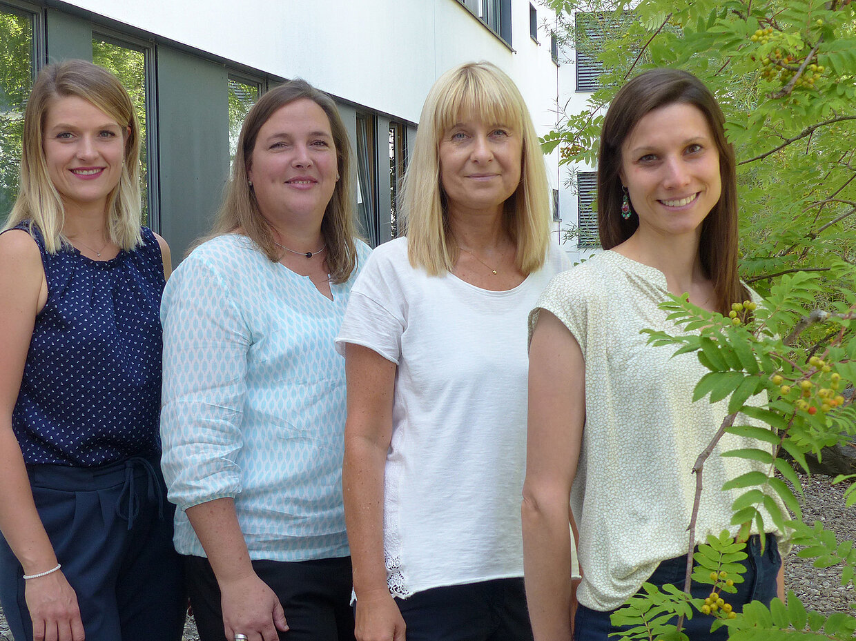 Vier Frauen stehen nebeneinander vor einem Gebäude mit großen Fenstern. Sie lächeln und tragen legere Kleidung in verschiedenen Farben. Einige Pflanzen sind im Vordergrund sichtbar, während der Hintergrund die Architektur des Gebäudes zeigt.
