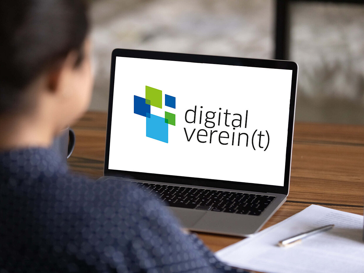 Eine Person sitzt mit dem Rücken zur Kamera vor einem Laptop, auf dessen Bildschirm das Logo "digital verein(t)" sichtbar ist. Der Hintergrund zeigt einen Holzschreibtisch und einen verschwommenen Bereich.