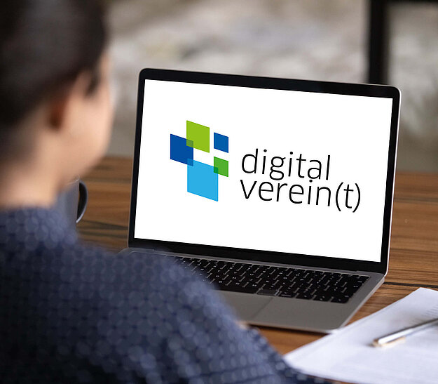 Eine Person sitzt mit dem Rücken zur Kamera vor einem Laptop, auf dessen Bildschirm das Logo "digital verein(t)" sichtbar ist. Der Hintergrund zeigt einen Holzschreibtisch und einen verschwommenen Bereich.