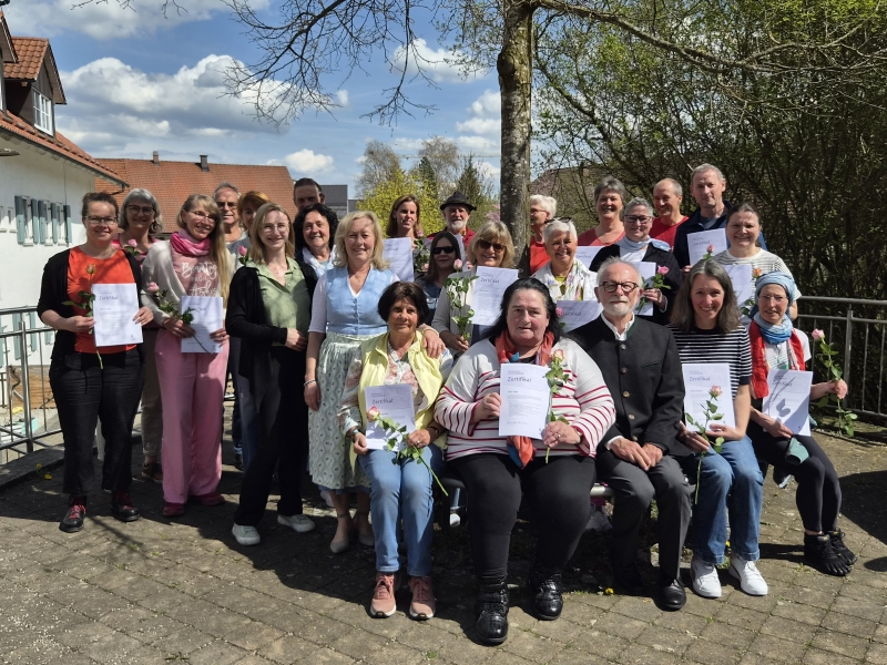 Eine Gruppe von Menschen steht auf einer Terrasse und hält Urkunden in den Händen. Sie lächeln in die Kamera. Im Hintergrund sind Bäume und ein blauer Himmel zu sehen. Einige Personen halten Blumen.
