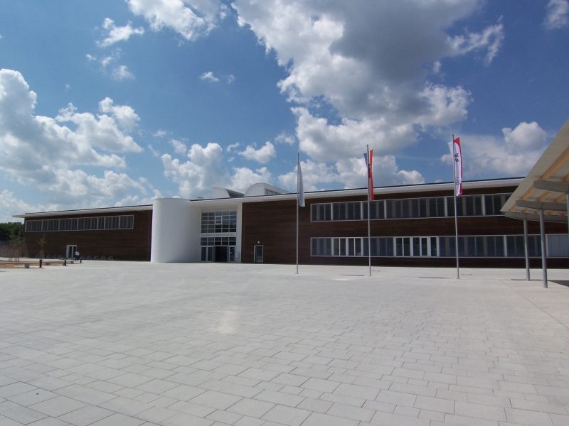 Bildbeschreibung: Ein modernes Gebäude mit großen Fenstern und einer Holzfassade, umgeben von einem gepflasterten Platz. Der Himmel ist klar mit einigen Wolken, und es stehen mehrere Fahnen vor dem Gebäude.