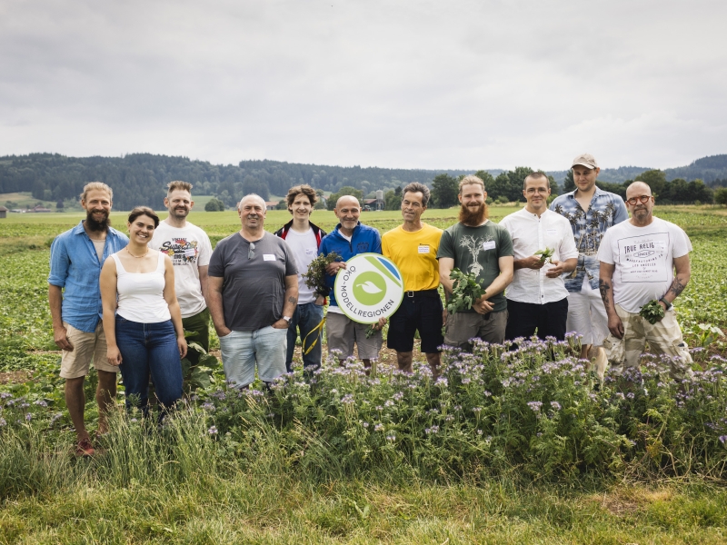 Eine Gruppe von zwölf Menschen steht auf einem Feld, umgeben von Pflanzen. Sie lächeln und halten einige frische Kräuter in der Hand. In der Mitte ist ein großes Schild mit einem grünen Logo zu sehen. Der Himmel ist bewölkt und die Landschaft erstreckt sich im Hintergrund.