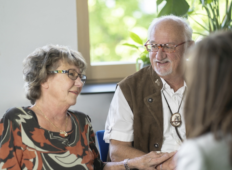 Ein älteres Paar sitzt zusammen in einem hellen Raum. Die Frau mit lockigem Haar trägt eine bunte Bluse und schaut den Mann lächelnd an. Der Mann hat ein freundliches Gesicht, trägt eine Weste und eine Kette. Hinter ihnen sind Pflanzen und Fenster zu sehen.