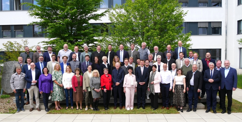 Gruppenfoto von etwa 50 Personen im Freien, vor einem modernen Gebäude und einer größeren Baumlandschaft. Die Teilnehmenden tragen überwiegend formelle Kleidung und stehen in verschiedenen Posen, einige lächeln in die Kamera.